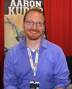 Posed photograph of a smiling, bearded Aaron Kuder in front of red curtained background at the 2012 New York Comic Convention, Kruder wears a blue shirt, no tie