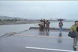 Scene from on the bridge post-collapse. Several reporters can be seen with umbrellas, and the weather is cloudy. The road is completely broken off and bent slightly upwards.