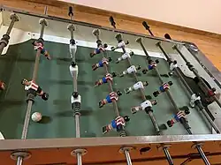 View of a game of table football. It consists of a table with holes on either end, for the goals, and with poles filled with miniature footballers and a ball for each player to control.