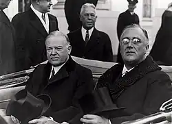 Hoover and Roosevelt sit in the back seat of an open top car with their hats on their laps.
