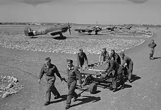 a black and white photograph of men pulling a trolley, with aircraft dispersed in the near background
