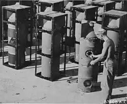 A black and white photograph of a shirtless man inspecting and loading multiple aerial mines