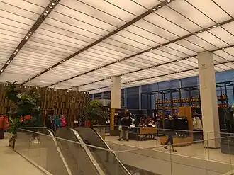 View of the second floor. There are racks of merchandise in the background and an escalator in the foreground. The ceiling is composed of a luminous material, and the lights are hidden behind the ceiling. The glass facade is visible at far right.