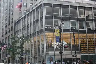 The facade of the building as seen from slightly further north on Fifth Avenue. The building is four stories high, with tall windows, vertical steel mullions, and horizontal black spandrel panels. The second story is significantly taller than the others.