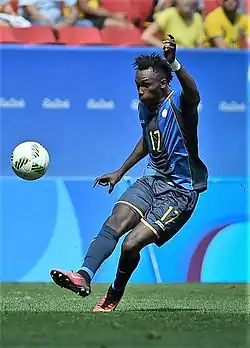 A man wearing navy blue shorts and a blue jersey kicking a football.