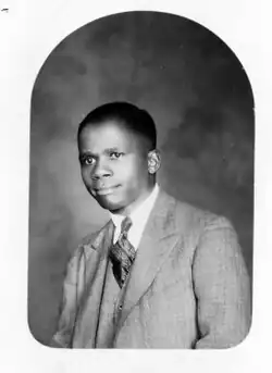 Black and white photo of a man in a suit facing forward towards a camera.