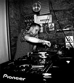 Andrew Weatherall, dressed in a black and white striped t-shirt, standing at a set of turntables while performing live