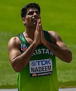 Arshad Nadeem looking up with his hands together in celebration. He is a man in his late 20s with short hair and no facial hair wearing a sports jersey.