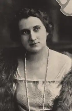 A woman with light skin and dark hair, wearing a light-colored gown, a strand of pearls, and a fur wrap