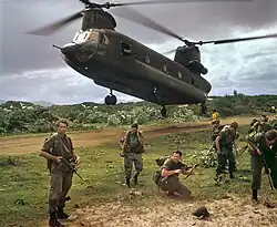 Colour photo of a group of men wearing military uniform and carrying guns standing in front of a twin-rotor helicopter which is flying just above the ground