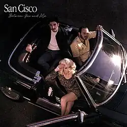 Two men and one woman sit in a retro car, looking up at the camera
