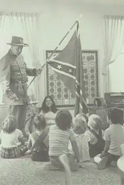 Black and white photo of man in confederate army uniform holding confederate flag in a classroom with children seated around him