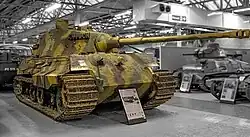 A large, turreted tank with dull yellow, green and brown wavy camouflage, on display inside Bovington museum. The tracks are wide, and the frontal armour is sloped. The long gun overhangs the bow by several meters.