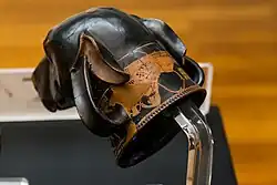 Photograph of the back of a Greek vase: the vase is shaped like a head of a dog. The dog has lean pointy ears that are folded backwards.