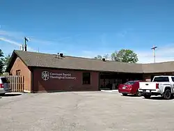 A section of a brick building with vehicles parked in front. On the brick wall is a sign reading "Covenant Baptist Theological Seminary" next to the seminary's seal.