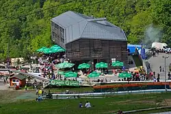 A hillside hotel in Pečovnik with a wooden structure and gray roof. Green trees make up the background and grass with reclining hotel guests in the foreground.