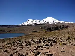 A barren, rock-strewn terrain with two ice-covered mountains in the background; to the left lies a blue lake and to the right a scarp.