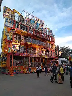 Photograph of a 5-story façade with open balconies. On top, lightbulb letters read "TORRENTE 5". Vertical surfaces are airbrushed with colorful realistic portraits and caricatures featuring Torrente and other characters.