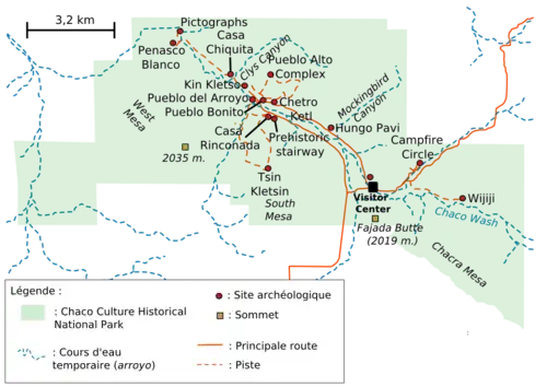 A large green area representing Chaco Culture National Historical Park's boundaries sits in the middle of a white field. The green area is roughly rectangular with one smaller square-like and one triangular appendage abutting it at bottom-left and bottom-right, respectively. Fifteen small red circles represent the location of important Chacoan sites; they are focused on a line running from top-left (northwest) to bottom-right (southeast). A dashed blue line depicting the Chaco Wash runs roughly along the same line; a network of dashed and solid orange lines represent trails and metalled roads, respectively, also focus on the same axis, connecting the red dots. Two gold squares define high points: "Fajada Butte (2019 m.)" and "West Mesa (2035 m.)".