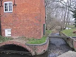 Close view of the south side of the building. A small eliptical brick arch leads the water into the enclosed millrace, and there is an overflow weir to the right of the building. Trees are visible on the riverbank beyond. The water is constrained in a red brick channel with a bulnose (curved) corner. Two scroll shaped iron 'plates' are on the upper wall, terminals for tie-rods that pass through the building. This is a winter view, and the state of the trees makes this stark. There is a small and unturbulent flow of water through the overflow weir, because the level is accurately controlled by the main weir in the old lock chamber. A precarious plank bridge, with no handrails, crosses the overflow race to reach the lockside on the right.
