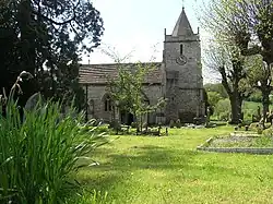 Gray stone building with small square tower and pyramidal roof. Grassy foreground with a cross and gravestones
