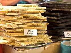 White chocolate bars piled imperfectly. Pieces of orange are visible within the bars. Behind them, dark bars appear stacked the same.