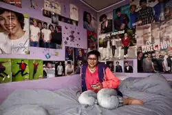 A teenage girl sits on a large bed with a grey blanket in her room, which has purple walls. The walls are adorned with posters of teenage boys.