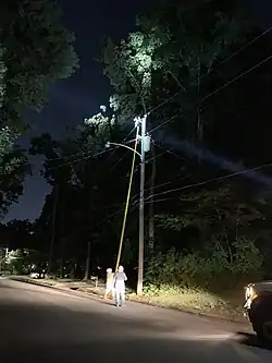 Two utility linemen with hard hats work to replace a cutout fuse 35 feet above them with a non-conductive pole with a hook on the top.