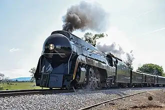 A black streamlined steam locomotive with a 4-8-4 wheel arrangement (four leading wheels, eight driving wheels, and four trailing wheels) and its tender, pulling an extra tender in the background