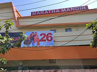 The side of a building with a sign that says "MARATHA MANDIR" and a billboard of the DDLJ poster with the words "Celebrating 26 years"