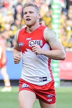A male athlete with light hair wearing a sleeveless guernsey and shorts jogs on the grass surface of the playing arena.