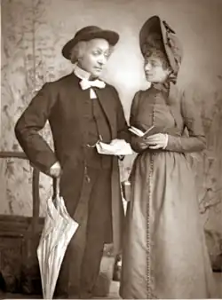 Middle aged white man, clean shaven, in clergyman's clothing with a young white woman in 19th-century costume