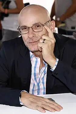 David Treuer wearing a light checkered shirt and dark blazer, sitting at a table, gazing directly at camera with chin propped up by left hand