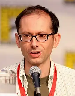 A closeup of a man in front of a microphone. He has a receding hairline and wears dark-framed glasses.