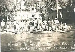 15 men lined up for a photo in and by a canoe, floating in the Río Fuerte, which has overflowed beyond its banks. Buildings are a couple meters behind them.