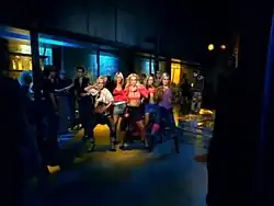 Image of five blond women. They are doing a choreographed dance in the middle of a dancefloor with the spotlight on them. Several people stand in the dark looking at them.