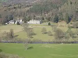 a country house in parkland between a river in the foreground and a forest in the background