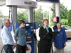 A white woman at a lectern on a Mobil gas station surrounded by another white woman and two white men.