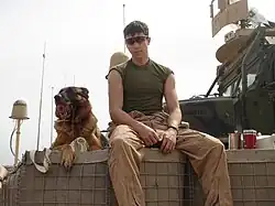 A black and tan German Shepherd dog sits next to a Marine atop a HECO barrier with a MTVR in the background. Lex is holding a red Kong toy in his mouth while Lee is wearing sunglasses and fire-resistant coveralls open to his waist.