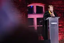 Else Baker standing at a podium in Auschwitz-Birkenau giving a speech