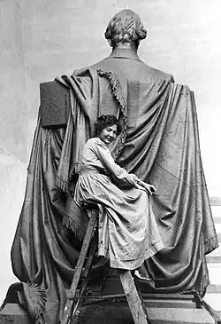 Sculptor Elsie Ward perched atop a ladder in a chamois work dress behind the sculpture which is in process