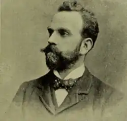 A vintage photograph of a man with a full beard and moustache, wearing a polka-dotted bow tie, a high-collared shirt, and a dark suit, looking to the side with a serious expression.
