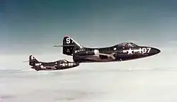 Two dark-blue-painted single-seat military jets flying from left to right in echelon. They wear the mark of the U.S. military on the nose, and a number. The nearer plane is 107 and the further is 116. On the fin is the letter 'S' and just in front the word NAVY. The planes have wingtip drop tanks and bubble canopies.