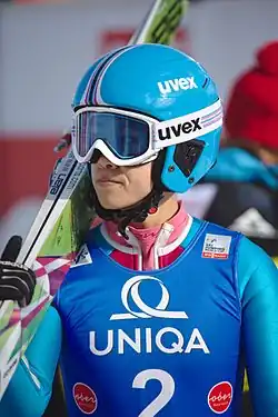 A professional skier wearing ski goggles. The skier is also wearing a blue helmet and holding skis over her shoulder.