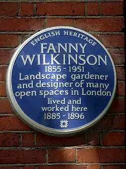 Round blue plaque with white text reading "English Heritage, Fanny Wilkinson 1855-1951 Landscape gardener and designer of many open spaces in London lived and worked here 1885-1896"