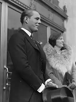 Two people standing in front of two sets of double doors, facing to the right. In front of the nearest set of doors is a man wearing a black suit with striped pants holding a top hat in front of him. In front of the far doors is an older woman wearing a fur-collared coat, clutching a purse in front of her with both hands.