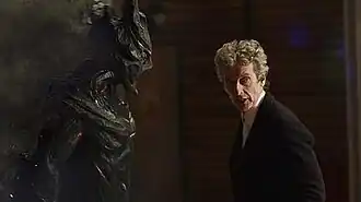 A screenshot from the episode featuring two actors from the series inside a school building, one has the appearance of an extraterrestrial creature