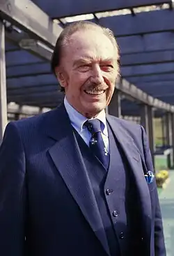 Photographic portrait of a balding blondish older man with a mustache. He is smiling, and his prominent eyebrows and lower eyelids nearly conceal his blue eyes. His right cheekbone is sunken in around the upper area of that side of the jawbone. His perfect teeth are just off-white. He is wearing a blue suit and tie.