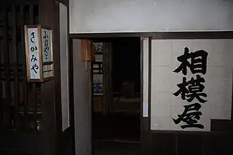 Dimly-lit narrow shopfront, with large black characters written in a single column on a shoji. A washi-and-frame lantern, mounted to protrude from the pillar, also bears signage.