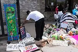 In front of a building, a sidewalk, raised curb, and nearer part of the street. From the sidewalk, over the cub, and onto the street, a pile of posters, bouquets of flowers, balloons, a plush seal toy, and a candle in a glass vessel. Draped over the top of the pile is an American flag in black and white. To the left of the pile, a man in the foreground faces away, bent over at the waist, holding up a framed picture portrait against a sidewalk fixture and the sidewalk, to the right of an artwork also leaned against the fixture. In the background to the right, a group of people.